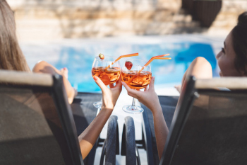 two women sitting in chairs by the pool clinking cocktail glasses together