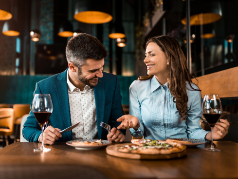 man and a woman sitting next to each other at a restaurant sharing pizza and wine